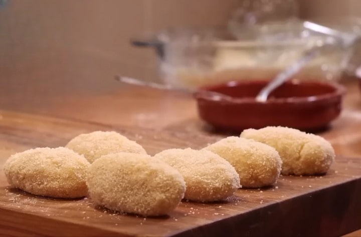 Croquetas de jamón y queso caseras formadas y rebozadas en pan rallado, listas para freír sobre una tabla de madera.