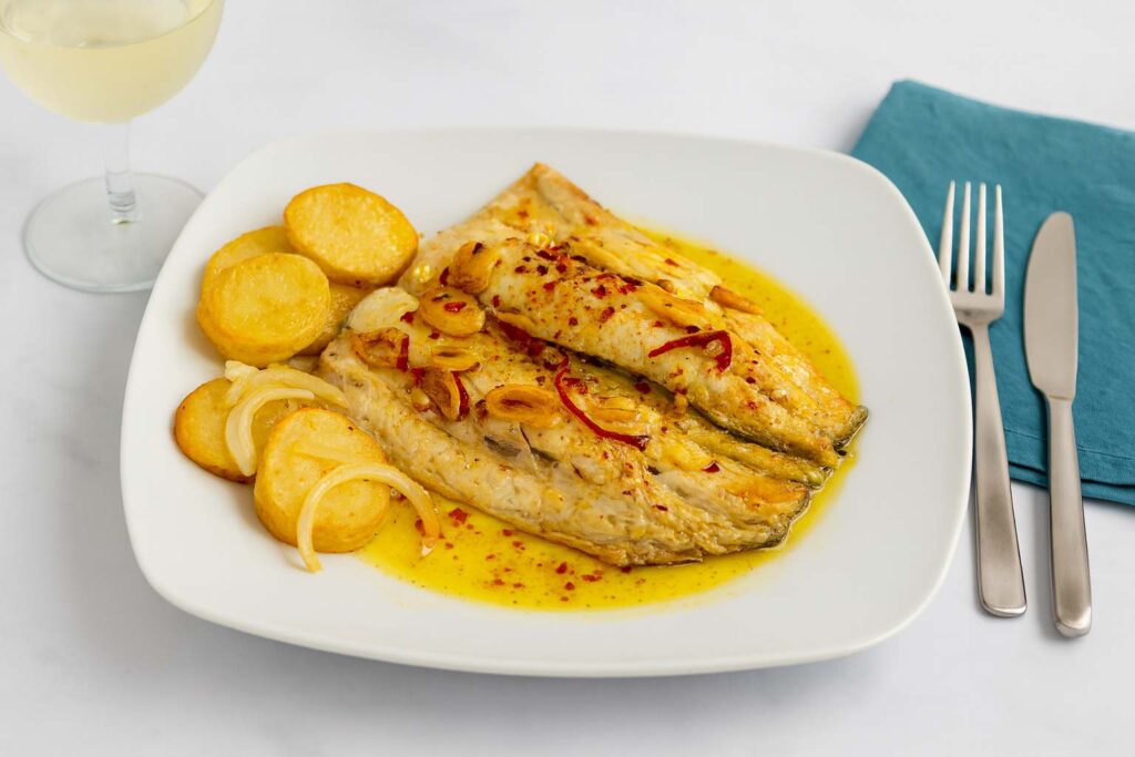 Vista de plato moderno con lubina al horno perfectamente hecha con sus patatas panaderas, cebolla y su refrito de ajo y guindilla listo para degustar